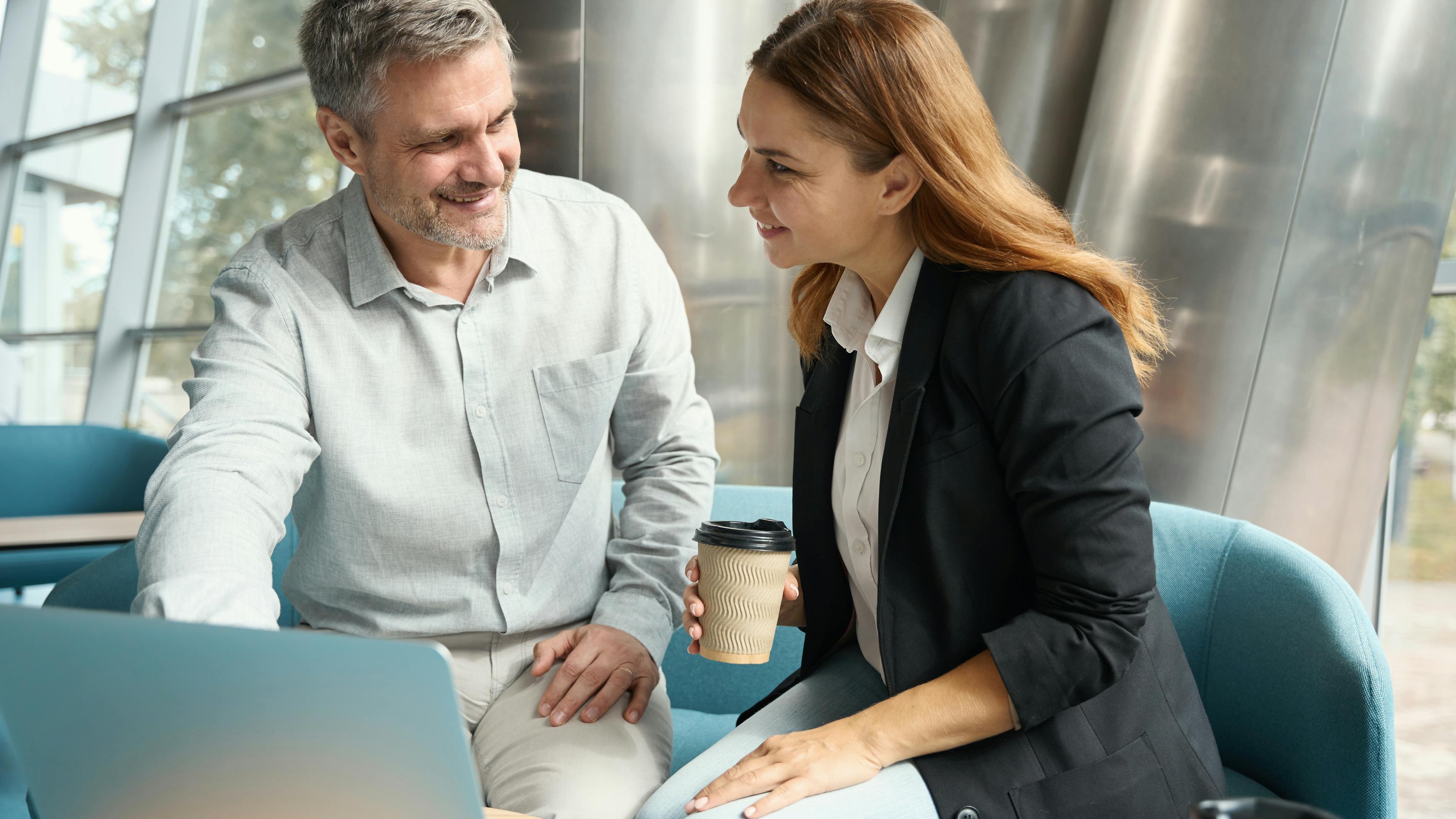 Two professionals in a contemporary office break area having a relaxed conversation, coffee cups in hand,