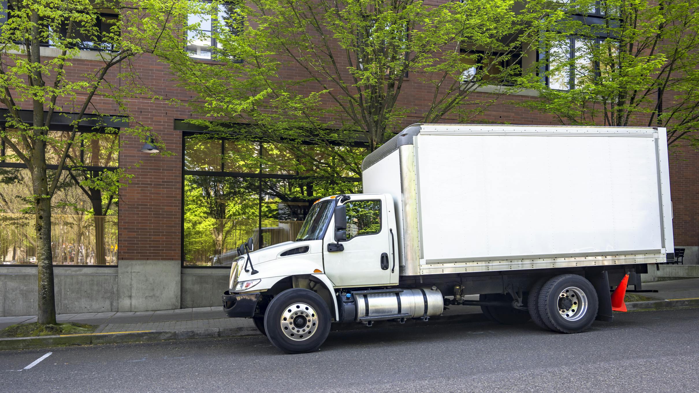 Middle duty day cab rig semi truck with box trailer make delivery standing on the city street with red bricks multilevel apartment.