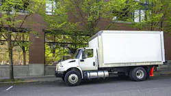 Middle duty day cab rig semi truck with box trailer make delivery standing on the city street with red bricks multilevel apartment.